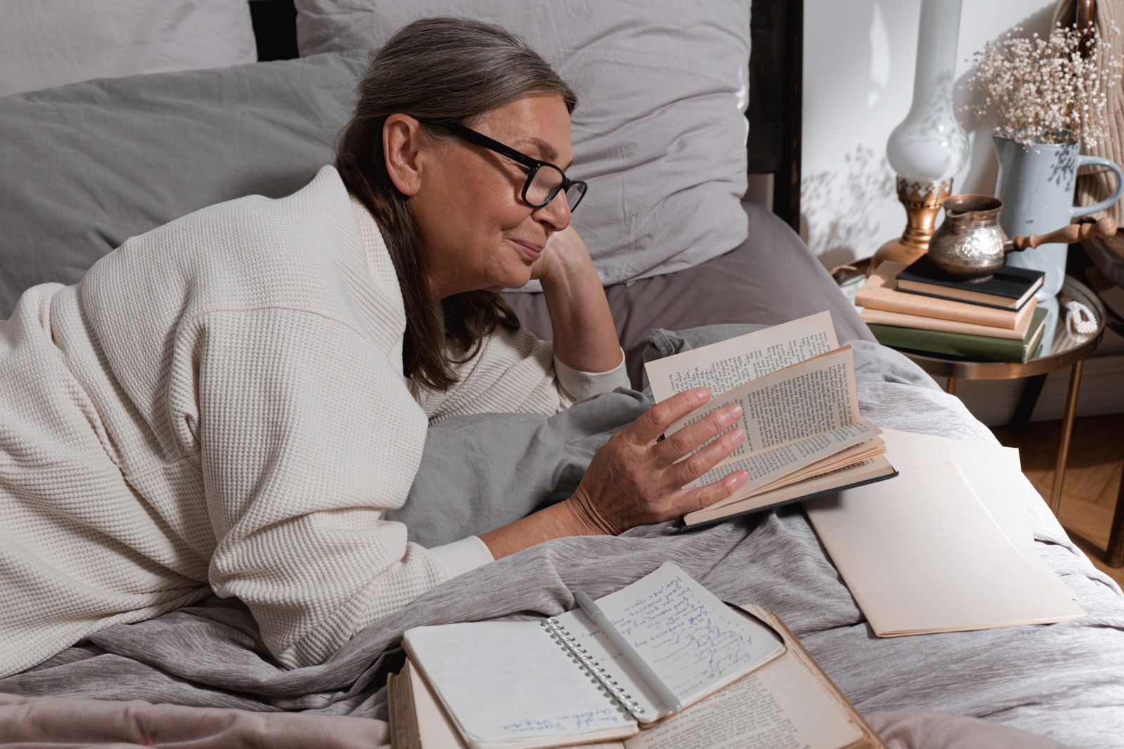 An older woman reading a book in bed at home, with a lamp and books on the nightstand