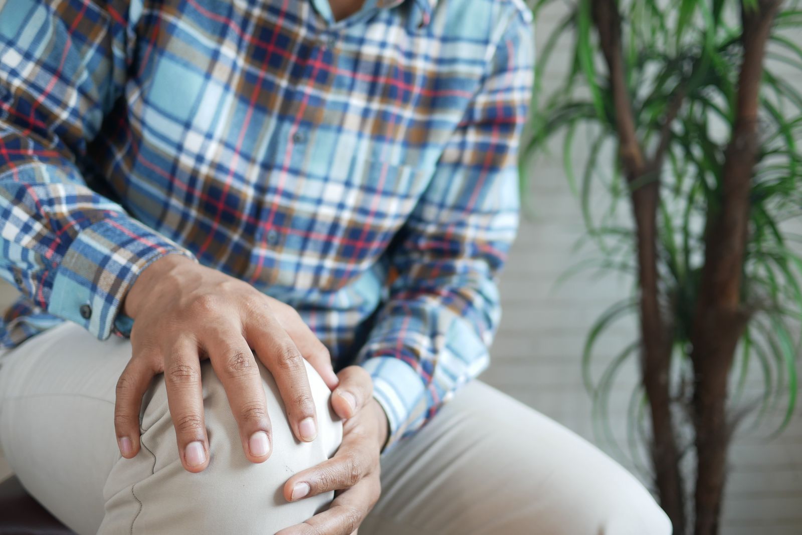 A seated man in a plaid shirt resting both hands on his knee indoors