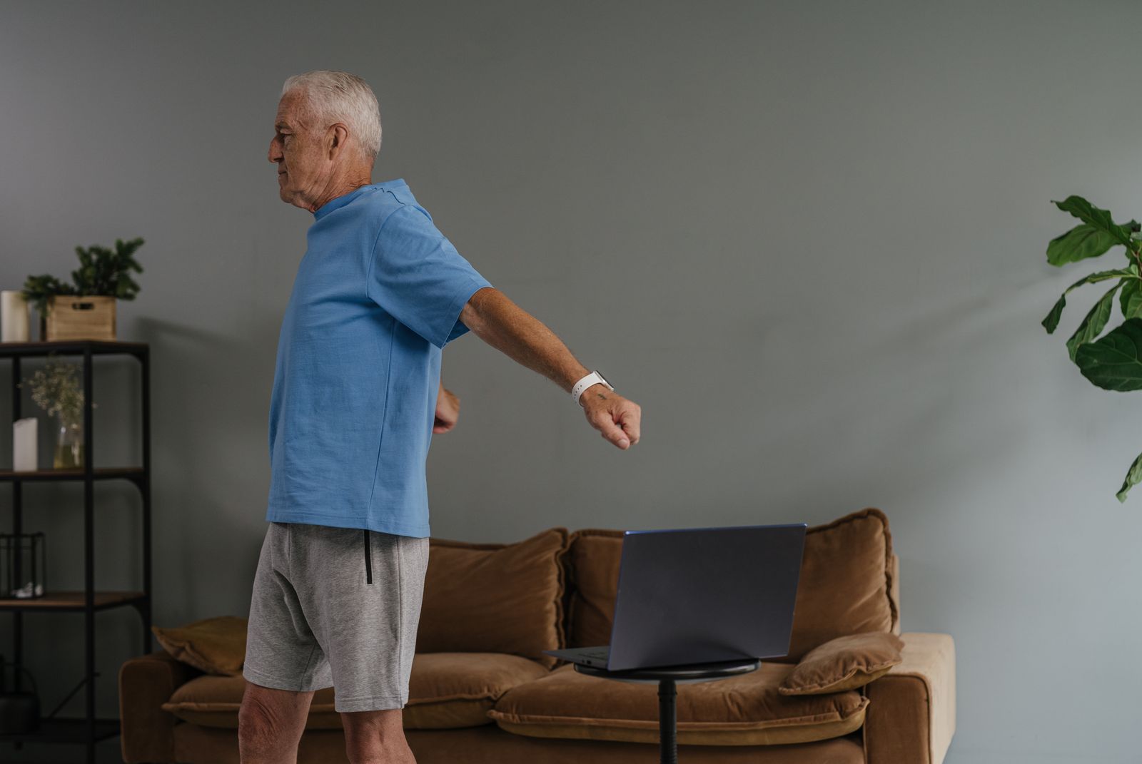 Older man in a blue shirt and gray shorts doing a standing arm exercise in his living room, a laptop open on a small table in front of the sofa