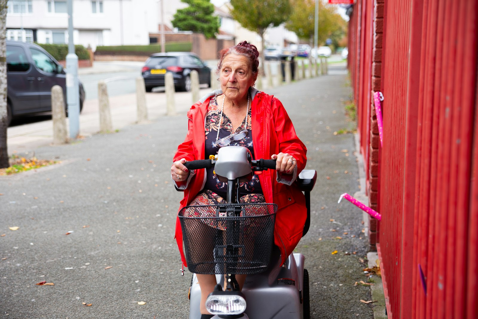 An older woman in a red coat riding a four-wheel mobility scooter along a sidewalk