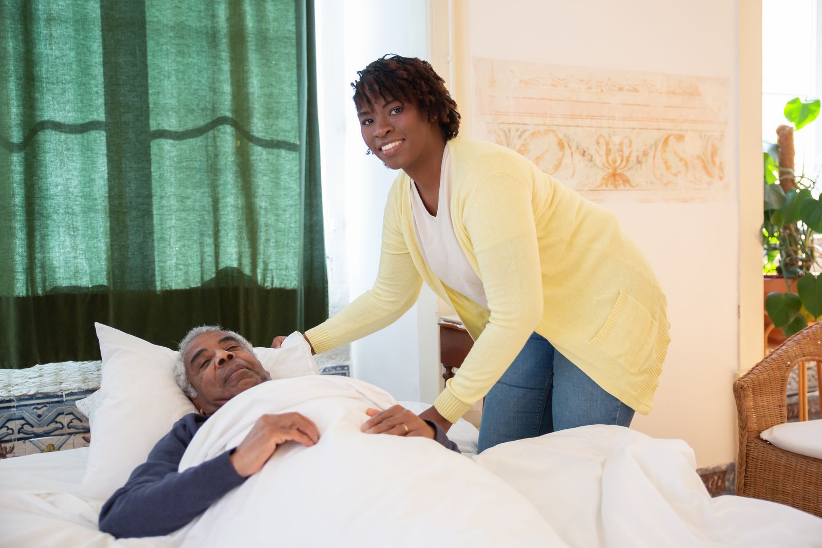A caregiver leaning over an older man in bed at home, adjusting his pillow before helping him sit up