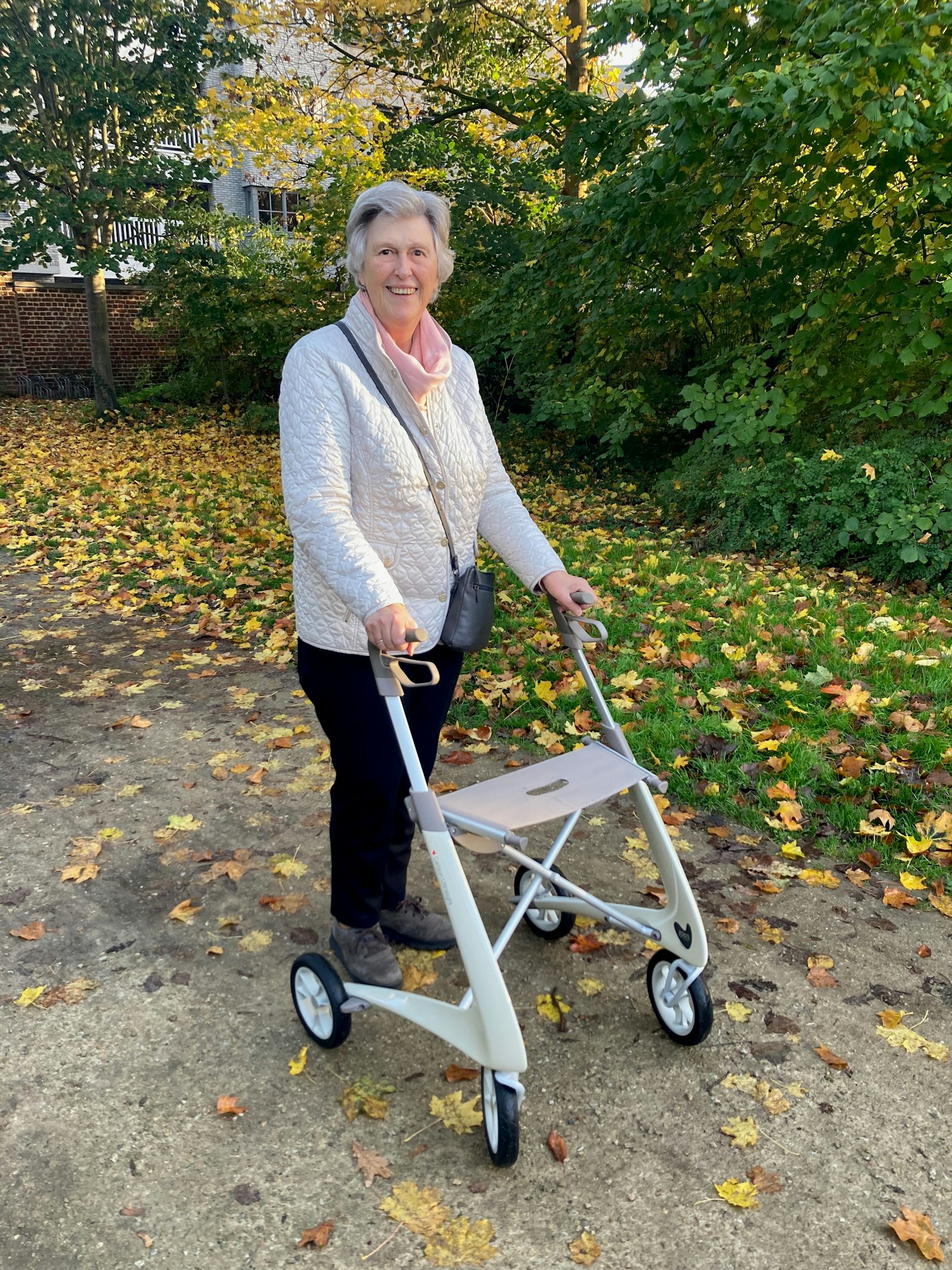An older woman walking a four-wheel rollator along a leaf-covered park path
