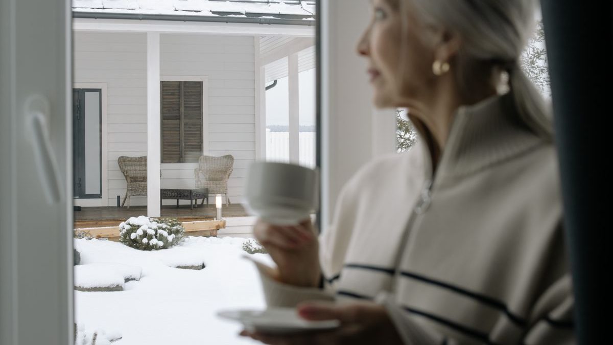 An older woman with a warm drink looking out at a snow-covered porch